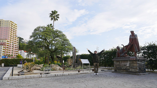 お宮の松と近くの銅像の間に立ちポーズを決めるライター地主恵亮